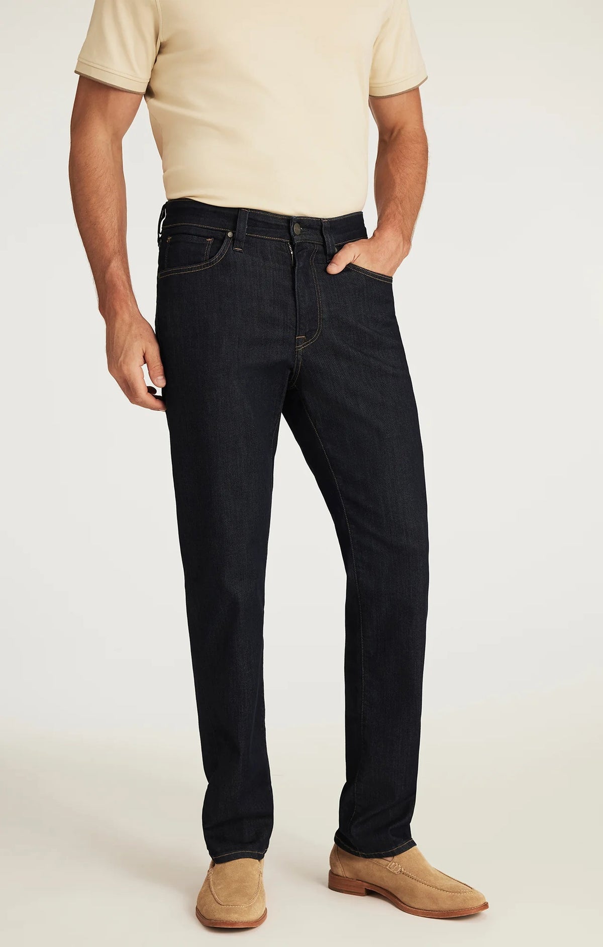 A man wearing a beige short-sleeve shirt, tan suede shoes, and the 34 HERITAGE - CHARISMA RELAXED STRAIGHT JEANS IN RINSE VINTAGE by 34 HERITAGE stands with one hand in his pocket against a white background.