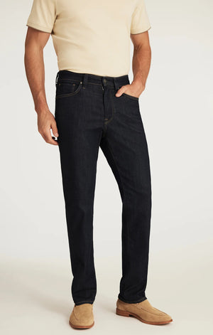 A man wearing a beige short-sleeve shirt, tan suede shoes, and the 34 HERITAGE - CHARISMA RELAXED STRAIGHT JEANS IN RINSE VINTAGE by 34 HERITAGE stands with one hand in his pocket against a white background.