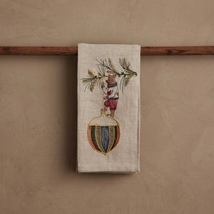 DECK THE HALLS BEAR LINEN TEA TOWEL featuring a bear in a sweater hanging a large, colorful, striped Christmas ornament from a pine branch on natural flax fiber.