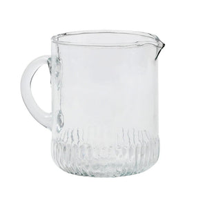 The ETUHOME - BISTRO GLASS MINI JUG by ETUHOME, filled with milk, rests on a white surface next to a butter dish and knife, with a clear glass baking dish and wooden container in the background.