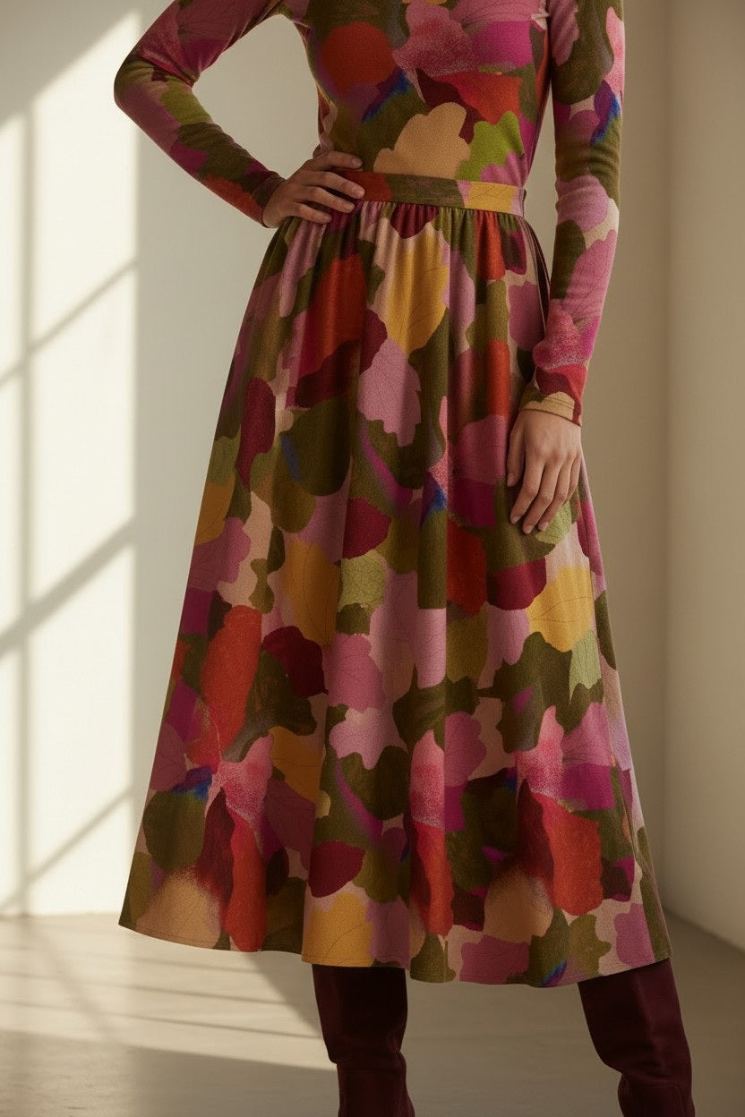 A person stands indoors near sunlight, dressed in a long-sleeved top paired with the FRANCO FERRARI - FRAGOLINA SKIRT by FRANCO FERRARI, which features a bold and colorful floral pattern in shades of pink, red, green, and yellow.