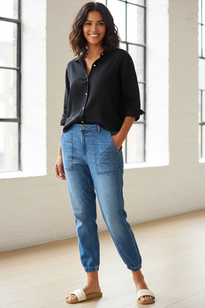 A person stands indoors with their back to the camera, wearing FRANK & EILEEN’s Jameson Utility Jogger Jean in 1968 Wash, a black top, and white sandals. A beige couch and patterned rug appear in the background.