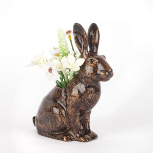 The QUAIL DESIGNS QUAIL CERAMICS - HARE BUD VASE is a hand-painted stoneware vase shaped like a rabbit, featuring a brown and black finish. It displays small white flowers and greenery in the opening on its back against a plain white background.