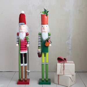 The CREATIVE COOP WOOD NUTCRACKER WITH FLOWER POT AND GARDEN TOOL wears green and yellow, holds flower pots as gardening tools, and sports a red flowerpot hat with a green sprout. Displayed on a white background.