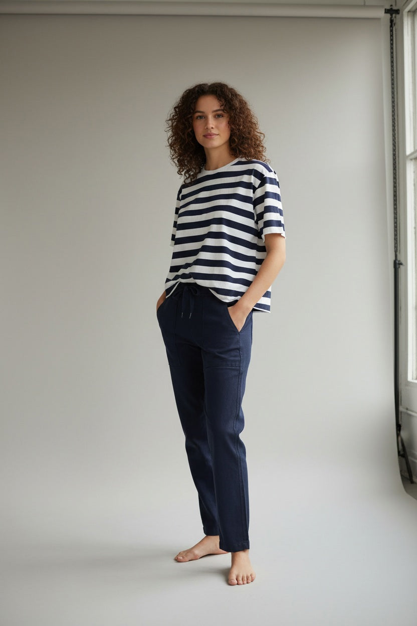 A woman stands outdoors in front of greenery, wearing the FRANK &amp; EILEEN PERRY NYC Crewneck Tee in Vintage Jersey Nautical Stripe and dark pants, with her hands in her pockets, smiling slightly at the camera.