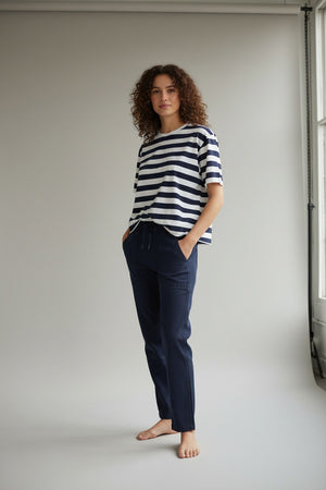 A woman stands outdoors in front of greenery, wearing the FRANK & EILEEN PERRY NYC Crewneck Tee in Vintage Jersey Nautical Stripe and dark pants, with her hands in her pockets, smiling slightly at the camera.