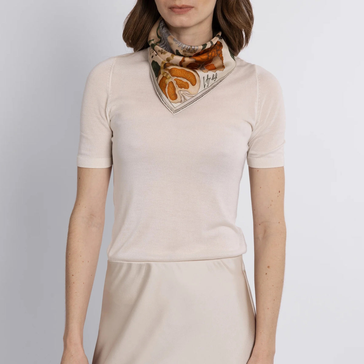 A woman in a beige short-sleeved top and matching skirt stands against a light background, adorned with the SABINA SAVAGE ORANGES AND LEMONS SILK TWILL SCARF in Cintrine/Zest. Her shoulder-length brown hair softly frames her face, enhancing her effortless elegance.