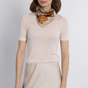 A woman in a beige short-sleeved top and matching skirt stands against a light background, adorned with the SABINA SAVAGE ORANGES AND LEMONS SILK TWILL SCARF in Cintrine/Zest. Her shoulder-length brown hair softly frames her face, enhancing her effortless elegance.