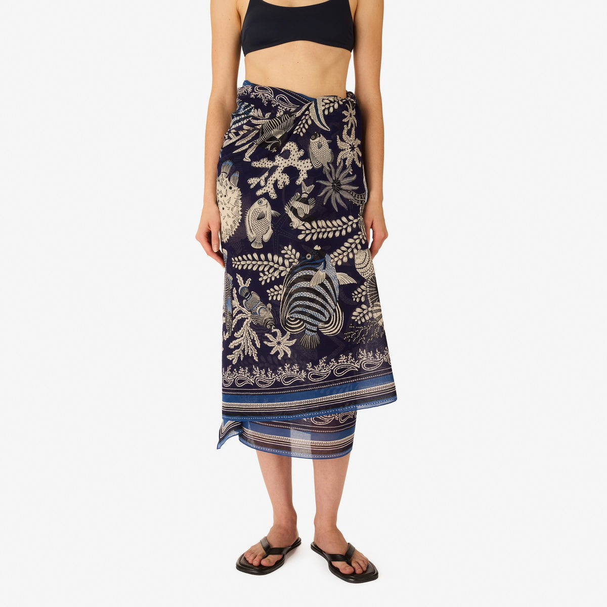 A woman faces forward, wearing a sleeveless cream and navy top with white botanical and animal motifs, accessorized with the INOUI EDITIONS - NEPTUNE SCARF IN NAVY by INOUI, draped loosely around her neck.