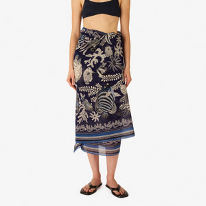 A woman faces forward, wearing a sleeveless cream and navy top with white botanical and animal motifs, accessorized with the INOUI EDITIONS - NEPTUNE SCARF IN NAVY by INOUI, draped loosely around her neck.