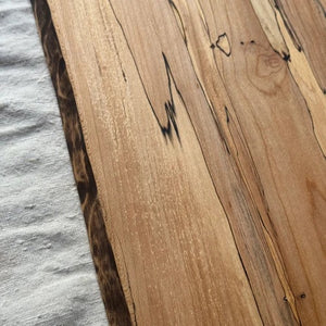 Handcrafted by PETERMANS BOARDS & BOWLS INC, the SPENCER PETERMAN - NATURAL EDGE SPALTED MAPLE RECTANGULAR BOARD 15" features a beautiful natural spalted maple grain, slightly angled sides, and a convenient small hole in the top right corner for hanging. Its dark rustic edges highlight the wood's unique character, making it an elegant choice for serving charcuterie or as a decorative cutting board.