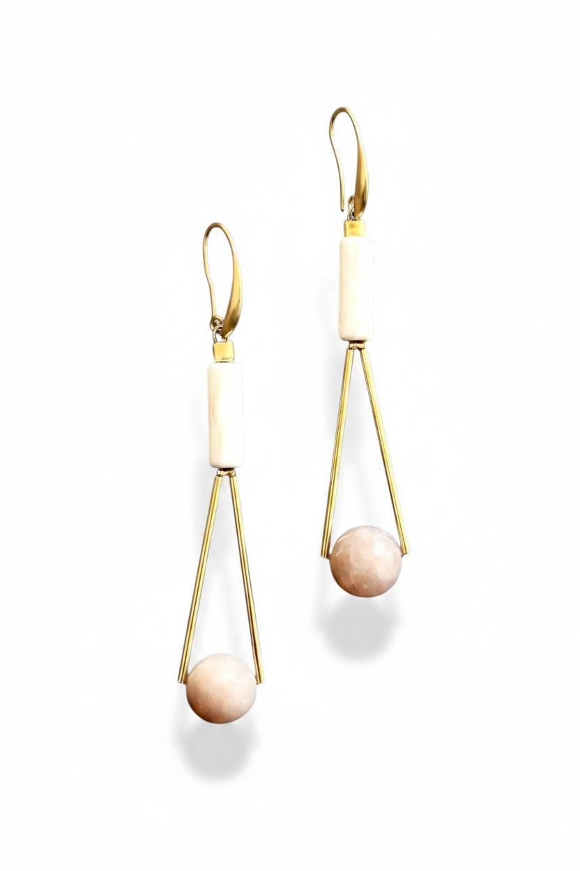 The DAVID AUBREY - PEACH MOONSTONE GEOMETRIC EARRINGS by DAVID AUBREY feature a sleek 18k gold plated triangular wire design, adorned with a white magnesite cylindrical bead at the top and a round peach moonstone bead at the bottom, showcased against a gray background.