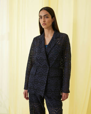A person with long, straight hair stands against a pale yellow background, donning the SFIZIO Double Breasted Broderi Anglaise Jacket by SFIZIO. The intricate circular patterns on the dark navy lace pantsuit add an elegant touch. The double-breasted jacket is buttoned up, and the person wears a serious expression with minimal accessories.