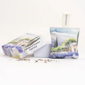A sachet from L'AUGUSTE PROVENCE, featuring a watercolor landscape and "Provence" fabric tag, stands upright on a white surface amid dried lavender. This French artisanal gift comes in a FRENCH ORGANIC LAVENDER SACHET AND GIFT BOX.