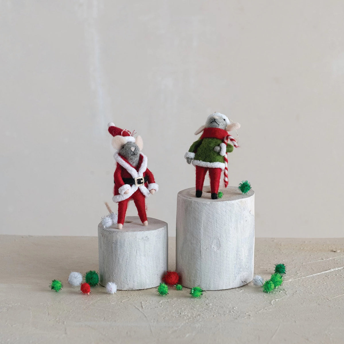The CREATIVE COOP FELT MOUSE ORNAMENT set includes two handmade wool felt mice in festive outfits, standing on white wooden blocks with red, green, and white pom-poms against a neutral, minimalist backdrop.