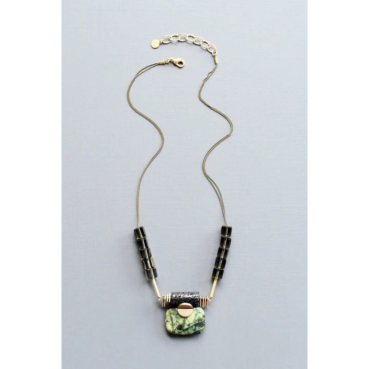 A close-up of the DAVID AUBREY Jasper ArtDeco Pendant Necklace by DAVID AUBREY, showcasing a marbled green jasper stone, a black lava-inspired textured bead, gold geometric accents, and a round gold element, all set against a gray background.