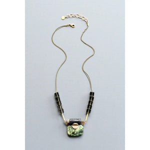 A close-up of the DAVID AUBREY Jasper ArtDeco Pendant Necklace by DAVID AUBREY, showcasing a marbled green jasper stone, a black lava-inspired textured bead, gold geometric accents, and a round gold element, all set against a gray background.