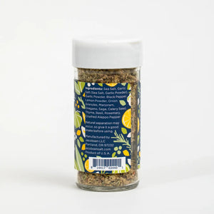 A jar of JACOBSEN SALT - ITALIAN SEASONING from JACOBSEN SALT CO. sits on a kitchen counter near utensils, a mason jar with a metal lid, and a ceramic cup. The spice jar label features a blue and yellow floral design.