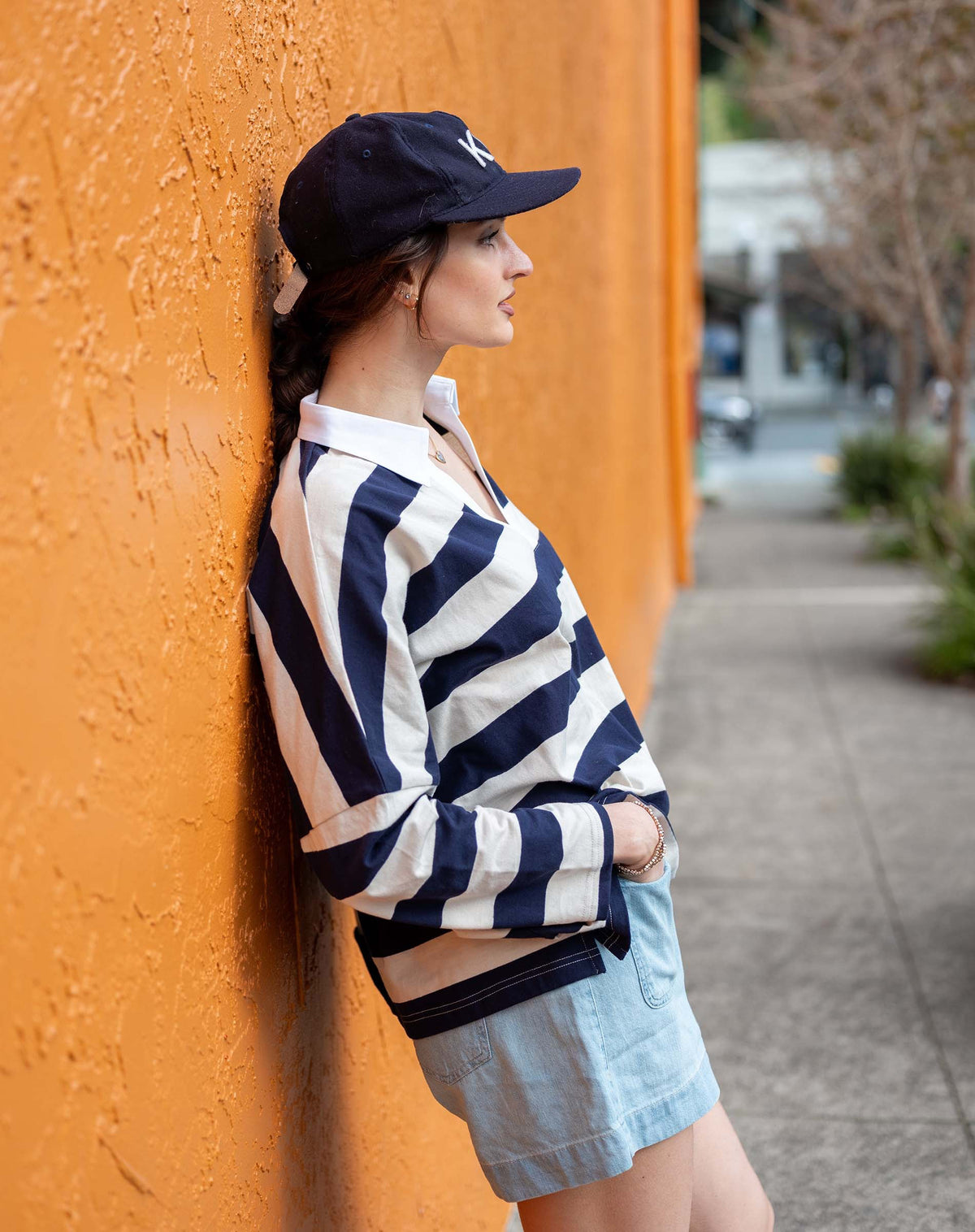 Wearing the MERSEA Amelia Rugby Tee in Navy Ecru Stripe, a woman pairs it with light denim shorts and a navy KC cap as she stands against an orange textured wall on a city sidewalk, gazing slightly to the side.