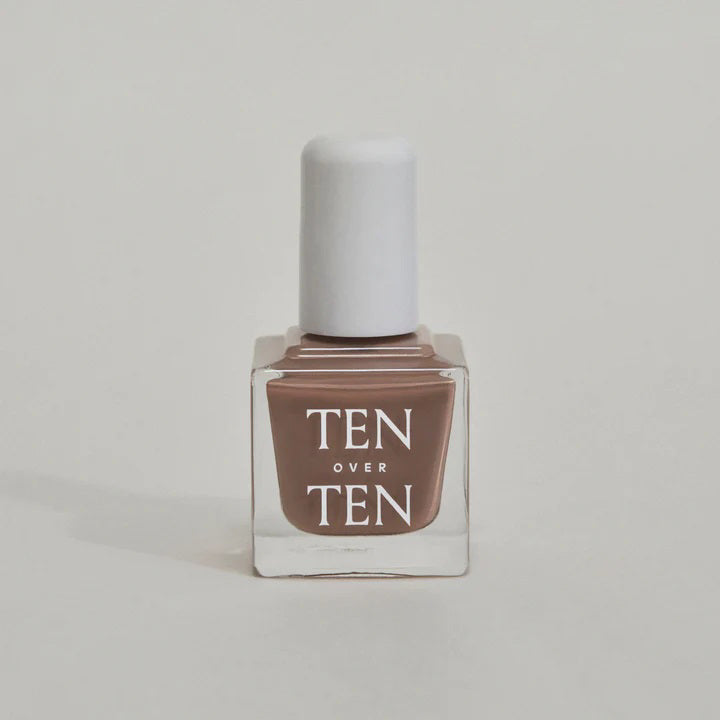 A glass bottle of TENOVERTEN - NAIL POLISH from the brand TEN OVER TEN, featuring a white cap and a dusty pink nail color. The front label displays "TEN OVER TEN" in white uppercase letters. This vegan, eight-free formula ensures both beauty and peace of mind. The background is plain and light-colored.