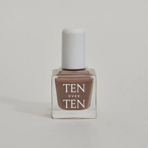 A glass bottle of TENOVERTEN - NAIL POLISH from the brand TEN OVER TEN, featuring a white cap and a dusty pink nail color. The front label displays "TEN OVER TEN" in white uppercase letters. This vegan, eight-free formula ensures both beauty and peace of mind. The background is plain and light-colored.