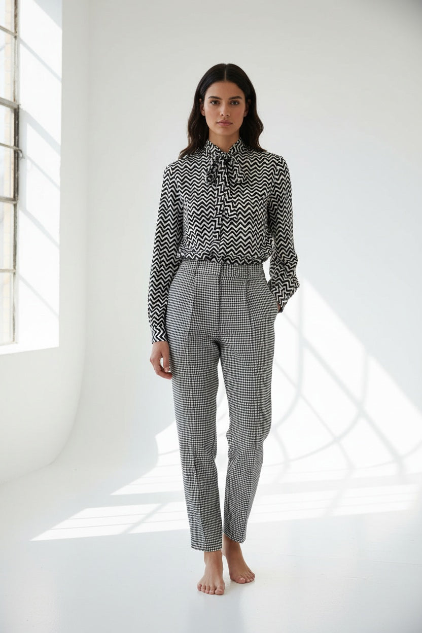 A woman with straight brown hair faces away in a HOPE FOR FLOWERS Tie Neck Blouse—black and white, vertically striped Tencel with puffed sleeves—paired with wide-leg plaid trousers. The plain gray background reflects sustainable style.