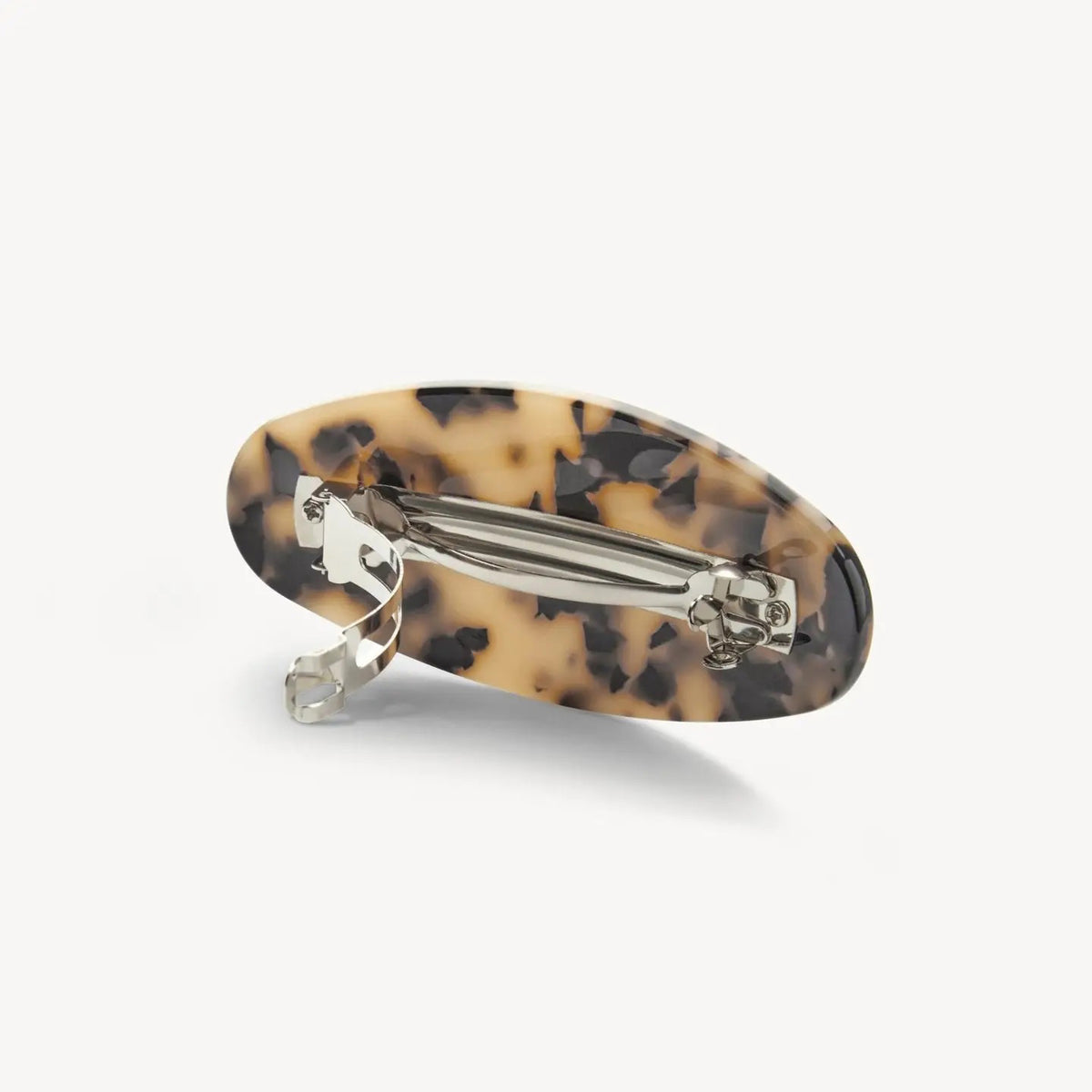 A person with wavy brown hair styled in a low twist, secured by the MACHETE Jumbo Oval Barrette in Blonde Tortoise, wears gold hoop earrings and a sleeveless black top, facing away from the camera against a white background.