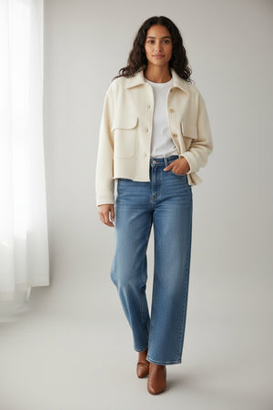 A woman with curly hair stands outside in a garden, wearing the FRANK & EILEEN Rowan Sweatshirt Utility Jacket in Triple Fleece Vintage White and blue jeans. A white house, greenery, and stone steps are visible in the background.