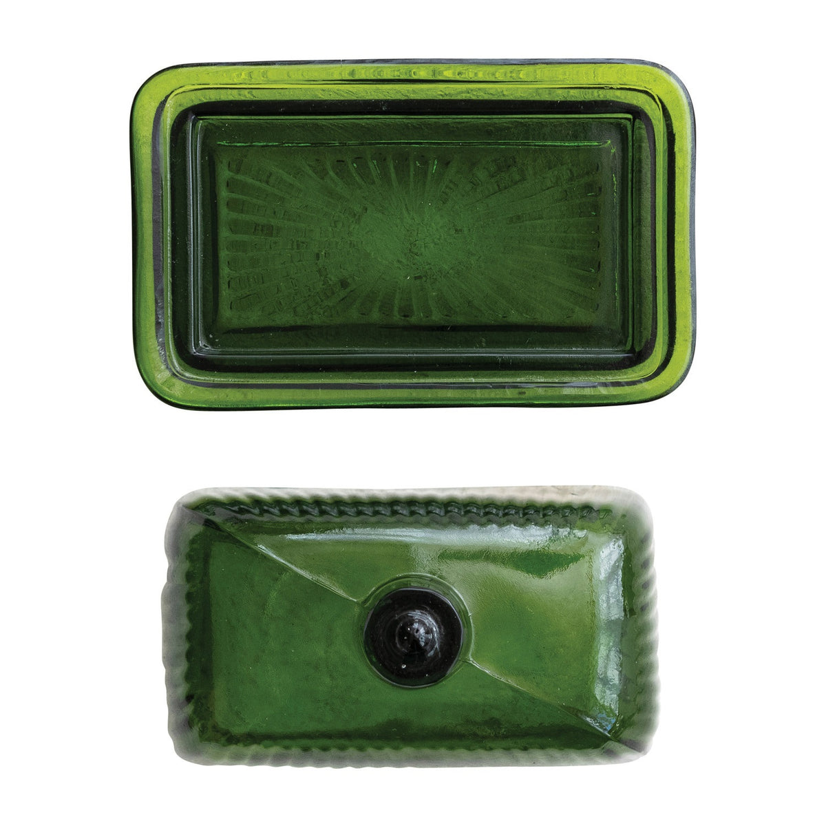 The CREATIVE COOP GREEN PRESSED GLASS BUTTER DISH is displayed with green glass containers and bottles on a textured white surface, beside a folded towel and a partially visible book.