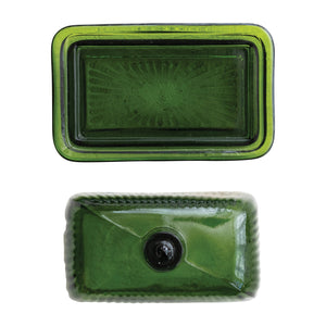 The CREATIVE COOP GREEN PRESSED GLASS BUTTER DISH is displayed with green glass containers and bottles on a textured white surface, beside a folded towel and a partially visible book.