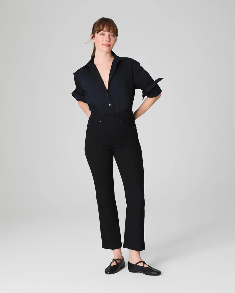 A woman stands against a light gray background, smiling with her hands behind her back. She wears the SPANX KICK FLARE JEAN IN CLASSIC BLACK, a high-waisted jean made from 4-way stretch denim, paired with a black button-up shirt and black flats.