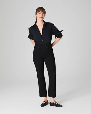 A woman stands against a light gray background, smiling with her hands behind her back. She wears the SPANX KICK FLARE JEAN IN CLASSIC BLACK, a high-waisted jean made from 4-way stretch denim, paired with a black button-up shirt and black flats.