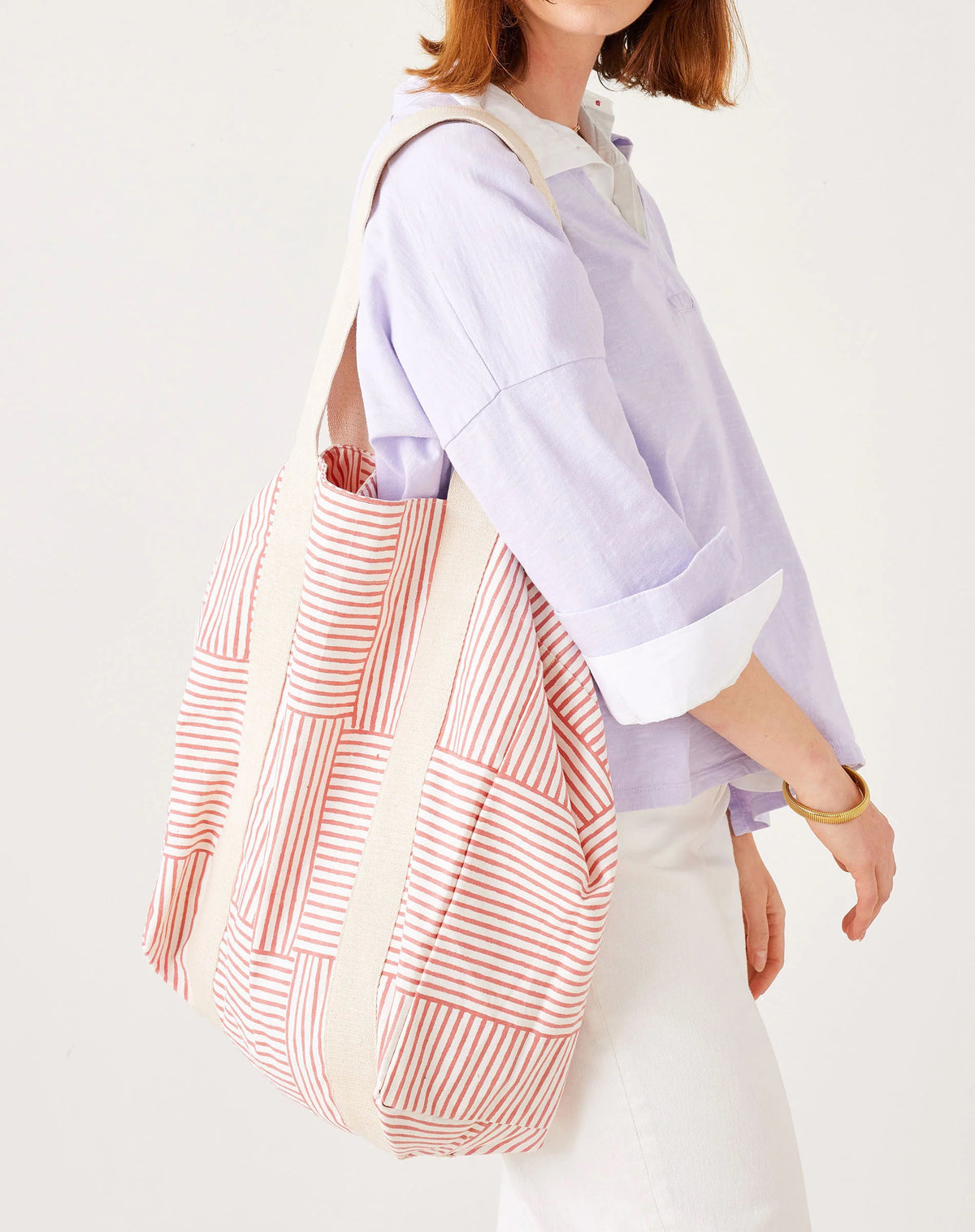 A woman in an orange long-sleeve shirt and denim shorts carries the MERSEA LEO TOTE BAG, a high-quality cotton tote from MERSEA with wide cream straps and an open top, featuring colorful orange, pink, and white checkered squares.