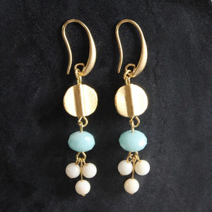 The DAVID AUBREY - AMAZONITE AND GLASS EARRINGS by DAVID AUBREY feature a round gold disk, a light blue amazonite bead, and three small white glass beads hanging from each earring, creating an elegant blend of classic style and contemporary charm against a striking black background.