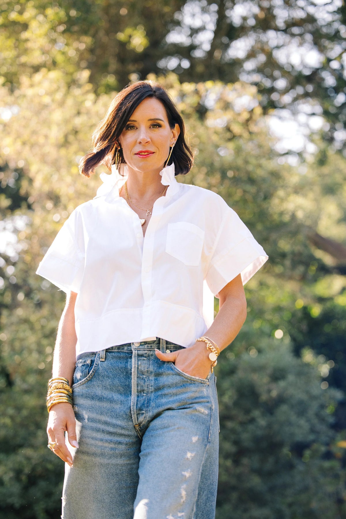 A woman in a black KMJ REESE SHIRT stands barefoot outdoors, leaning against a wooden post with one hand on her hip. She poses confidently, surrounded by lush green plants and trees.