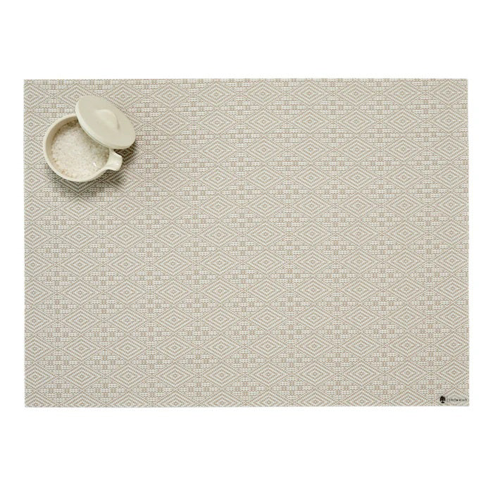 The CHILEWICH - BOXWOOD TABLE MAT by CHILEWICH in beige, featuring a subtle geometric diamond dobby weave, is styled with a small white salt bowl and lid positioned at the upper left corner.