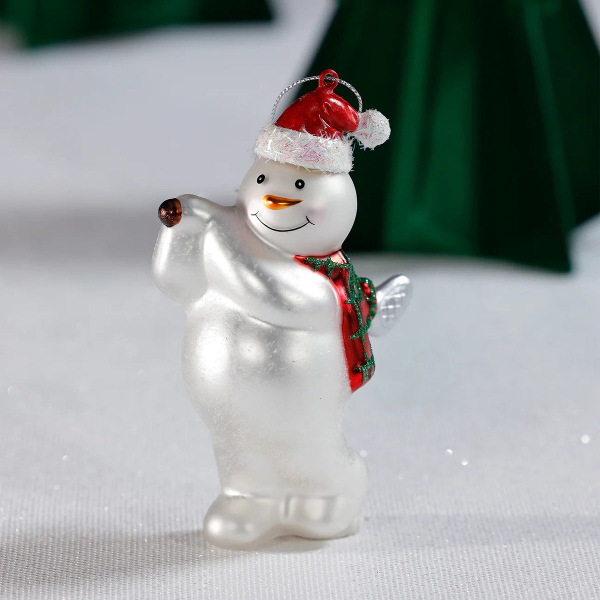 The ZODAX GLASS SNOWMAN PLAYING GOLF ornament, 3.5&quot;x5.25&quot;, hangs by a silver string and features a smiling snowman in a red Santa hat and green scarf, holding one arm up as he plays golf.