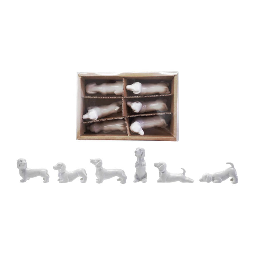 Six small white STONEWARE DOGS by CREATIVE COOP, each in a unique pose, are displayed in front of a six-compartment box, with each compartment holding a decorative dog figurine.