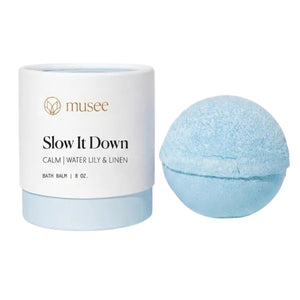 A handcrafted blue bath bomb rests beside its white and light blue cylindrical container labeled “MUSEE - SLOW IT DOWN THERAPY BATH BALM” from Musee.