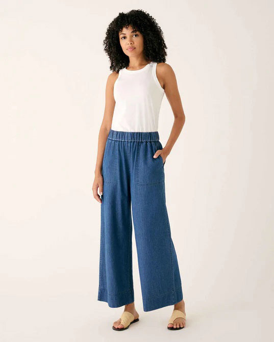 Wearing MERSEA - SAMMIE WIDE LEG PANT in light blue chambray with a pull-on waistband and front patch pockets, styled with a light top and cream sneakers, the person stands against a plain background.