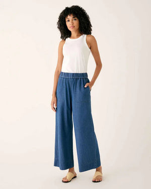 Wearing MERSEA - SAMMIE WIDE LEG PANT in light blue chambray with a pull-on waistband and front patch pockets, styled with a light top and cream sneakers, the person stands against a plain background.