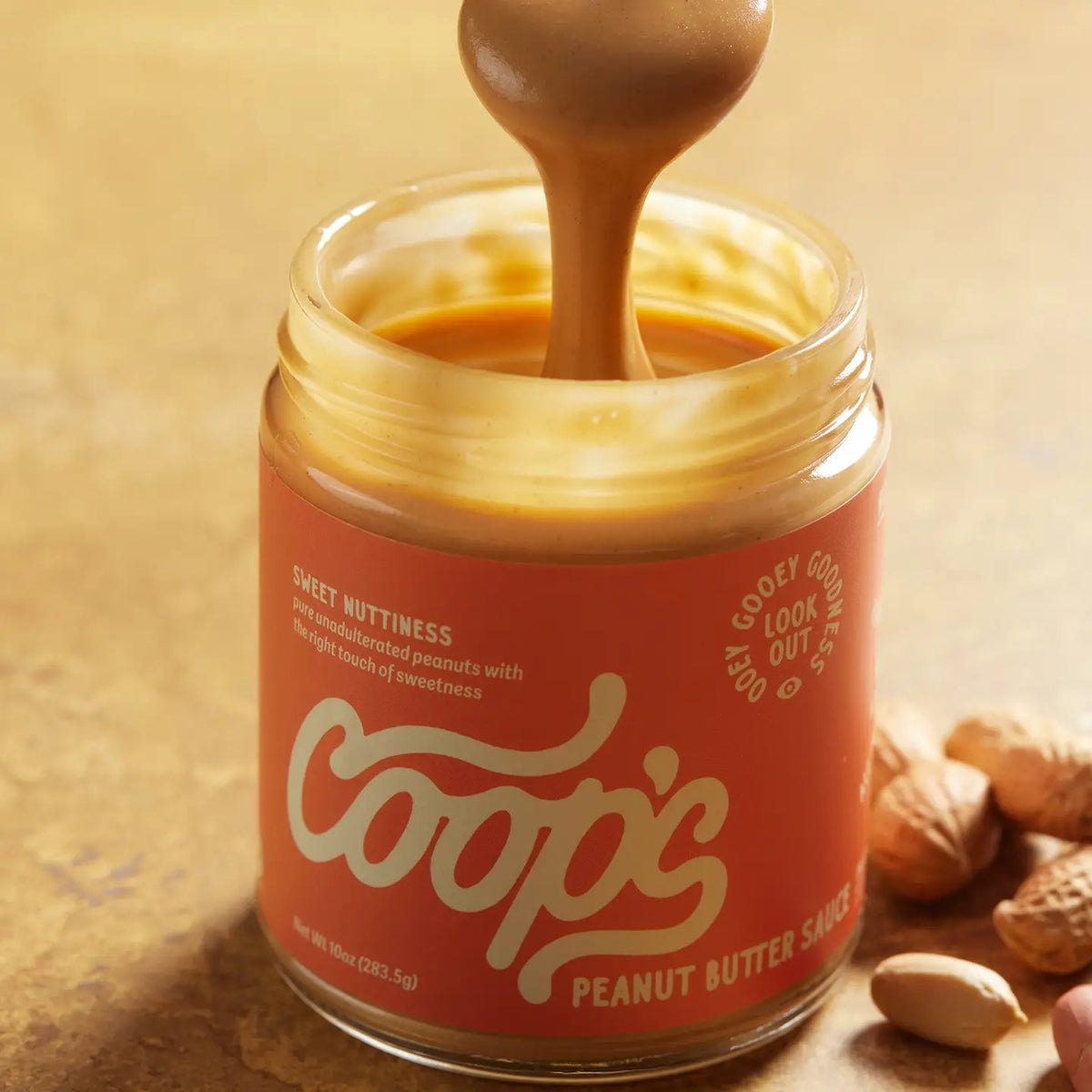 A 10 oz (283.5 g) jar of COOPS Peanut Butter Sauce, handmade in small batches to perfection, features chocolate-like drips down the lid and an orange label with “Sweet Nuttiness” and “Peanut Butter Sauce.”.