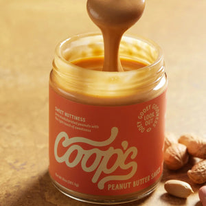 A 10 oz (283.5 g) jar of COOPS Peanut Butter Sauce, handmade in small batches to perfection, features chocolate-like drips down the lid and an orange label with “Sweet Nuttiness” and “Peanut Butter Sauce.”.