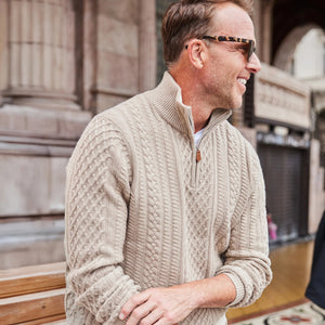 A man wearing sunglasses, white pants, and a COLLARS AND CO NAPOLI Cable Knit Quarter Zip Wool and Cashmere Sweater in Oatmeal holds a navy jacket over his shoulder while walking outside a historic stone building.