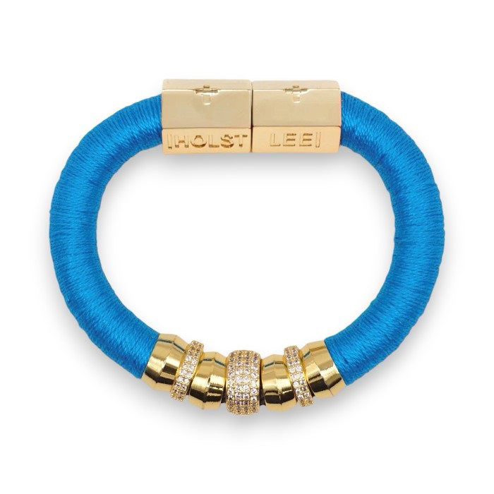 The HOLST + LEE - LUXE CLASSIC BRACELET by HOLST &amp; LEE features an elegant orange cord with gold-plated accents, including a rectangular gold clasp adorned with several gold and diamond-encrusted beads near the clasp.