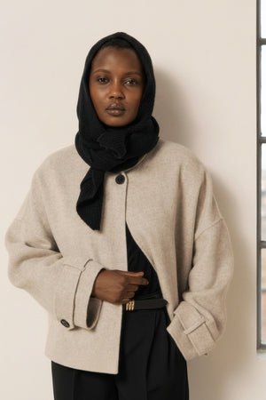 Wearing the HARTFORD - MERA Knit Hood/Scarf in Black—crafted from luxurious wool and cashmere—this person pairs a light long coat with a black turtleneck, wide-leg black pants, and gloves. They stand indoors against a light wall and wooden panel, striking a pose with one hand resting on their face.