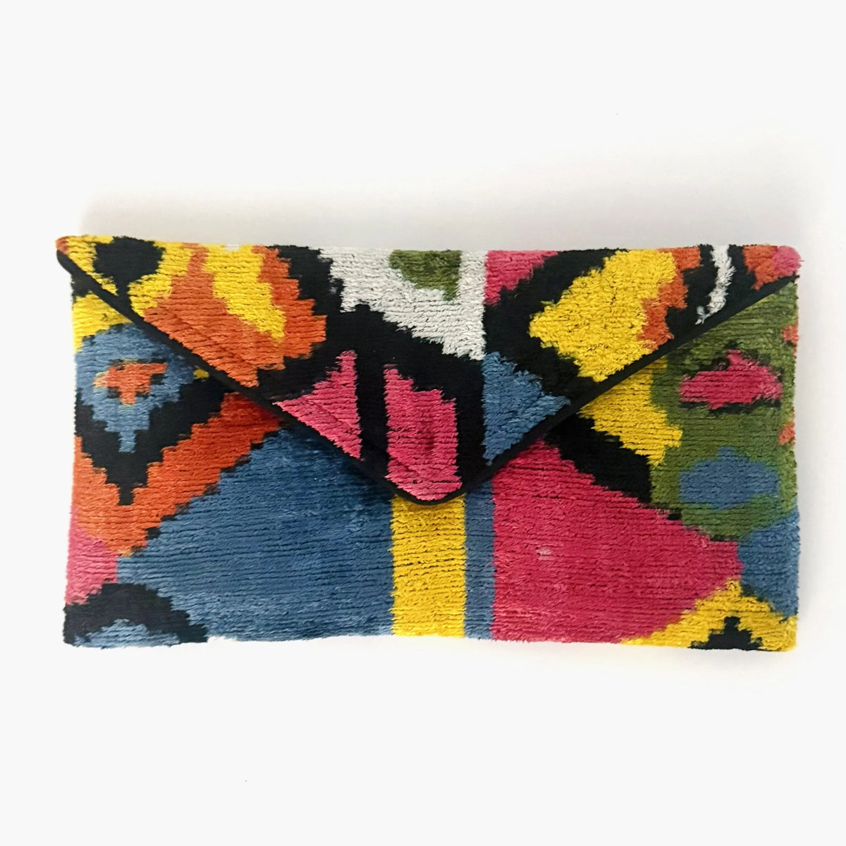 A person in a white outfit holds the BALDIZ - LILY CLUTCH IN PALOMA by BALDIZ, featuring a colorful geometric pattern. Their blue nails and a small forearm tattoo are visible.