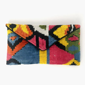 A person in a white outfit holds the BALDIZ - LILY CLUTCH IN PALOMA by BALDIZ, featuring a colorful geometric pattern. Their blue nails and a small forearm tattoo are visible.
