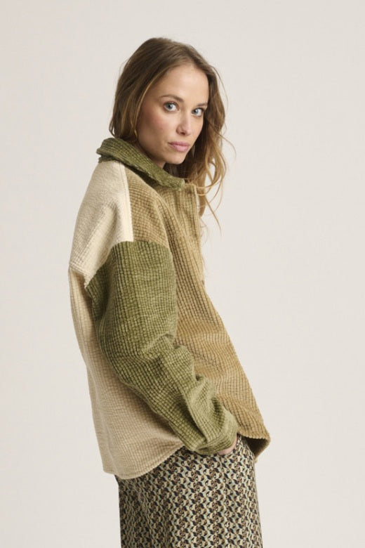 A woman with light brown hair wears the STORIATIPIC Corduroy Multi Shacket by STORIATIPIC—a tan and olive green button-up jacket crafted from soft cotton corduroy with a single chest pocket—paired with patterned pants. She stands against a plain light background, gazing at the camera with a neutral expression.