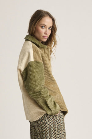 A woman with light brown hair wears the STORIATIPIC Corduroy Multi Shacket by STORIATIPIC—a tan and olive green button-up jacket crafted from soft cotton corduroy with a single chest pocket—paired with patterned pants. She stands against a plain light background, gazing at the camera with a neutral expression.